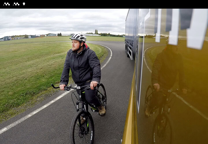 Cycliste portant un casque et une veste noire, roulant à côté d’un véhicule de livraison jaune sur une route rurale, illustrant la logistique et la gestion de flotte avec Microlise.