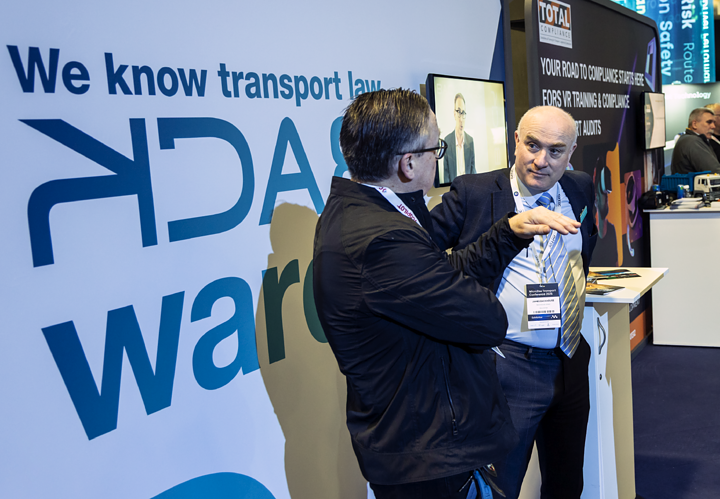 Two professionals engaged in a conversation at a Microlise event, focusing on fleet compliance and transport law, with branding and informational displays in the background.