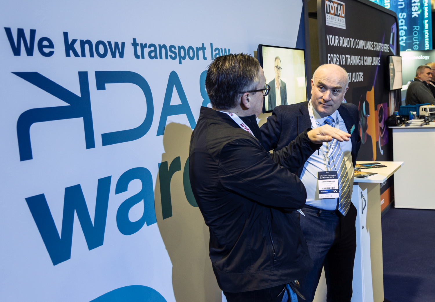Two professionals engaged in a conversation at a Microlise event, focusing on fleet compliance and transport law, with branding and informational displays in the background.