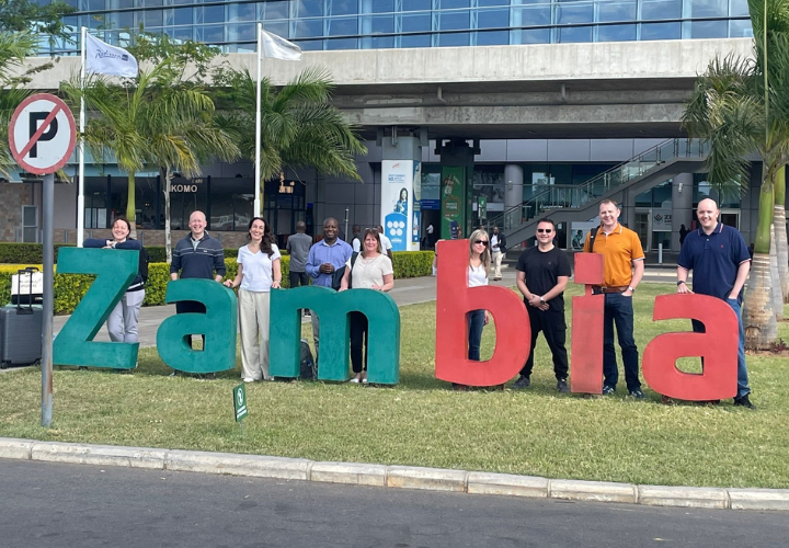 Zambia sign with diverse group of people standing in front, representing multicultural collaboration and community engagement at Microlise event or corporate gathering.