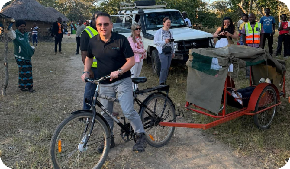 Man with sunglasses standing next to a bicycle with a trailer, outdoor event with people and vehicles in the background, rural setting.
